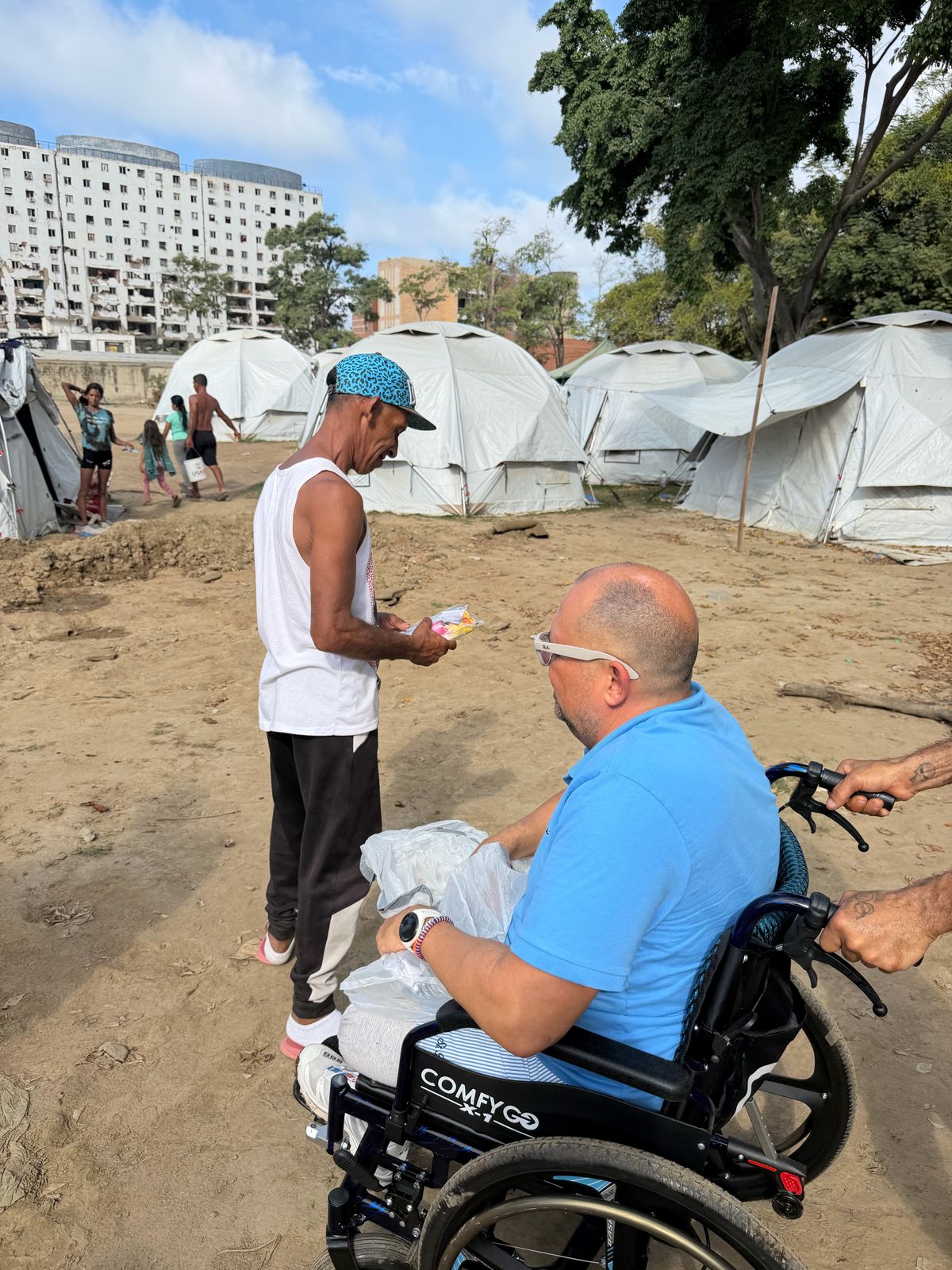 Un hombre en silla de ruedas con bolsas de suministros habla con un joven de gorra en un campamento de carpas blancas.