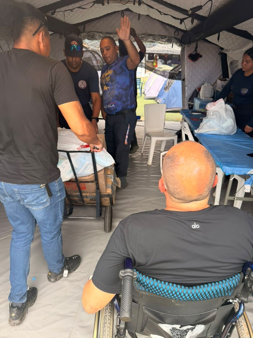 Visto de espaldas, un hombre en silla de ruedas frente a una carpa donde varias personas con uniforme reciben suministros.