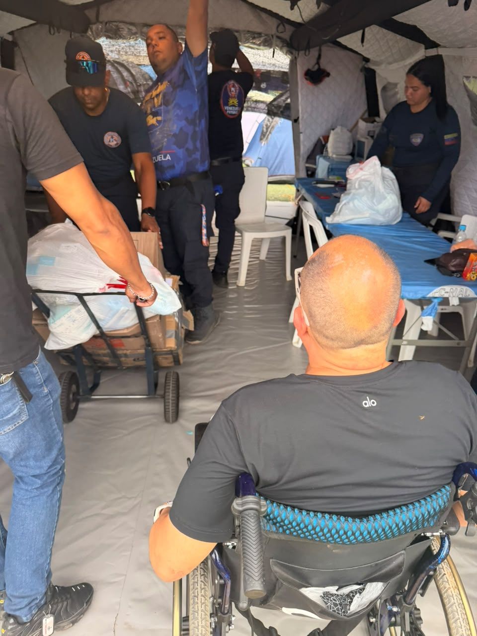Visto de espaldas, un hombre en silla de ruedas dentro de una carpa mientras varias personas, algunas con uniforme, mueven bolsas y suministros.