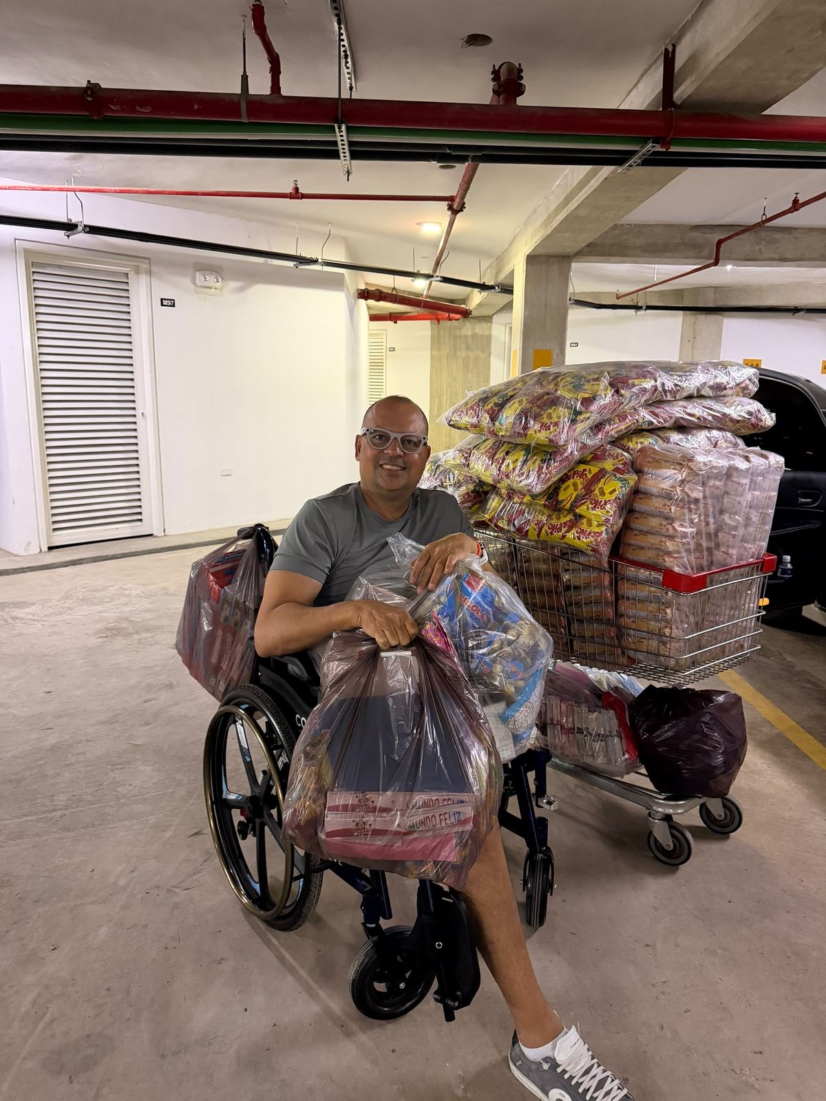 Hombre en silla de ruedas sostiene bolsas de suministros; a su lado, carritos cargados con paquetes de alimentos en un estacionamiento.