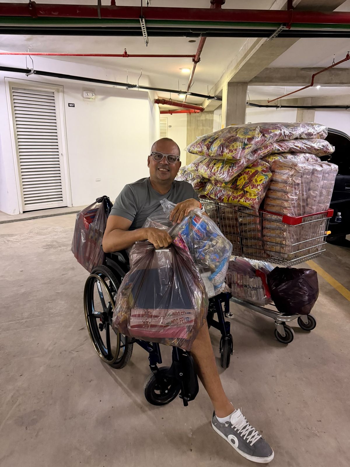 Hombre en silla de ruedas con bolsas de suministros en las piernas, frente a un carrito cargado con paquetes de alimentos en un estacionamiento techado.