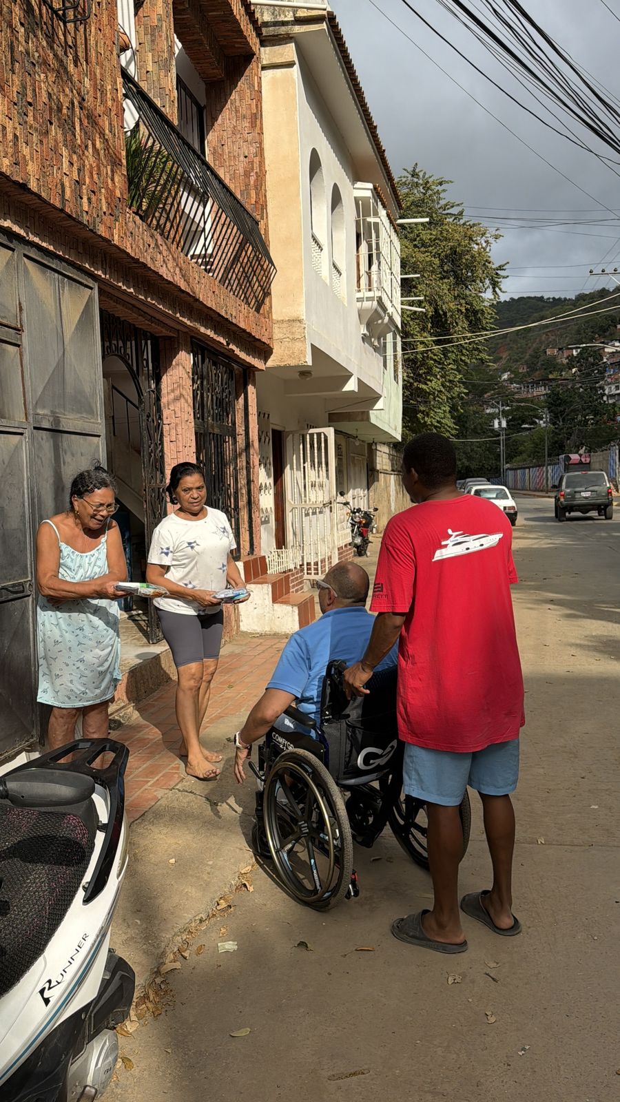 Un hombre en silla de ruedas conversa con vecinos en una calle estrecha entre casas.