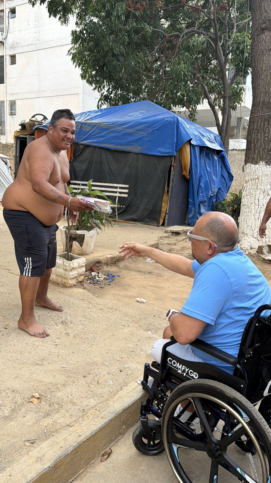 Un hombre en silla de ruedas extiende un paquete de ayuda a un hombre de pie frente a una carpa azul.