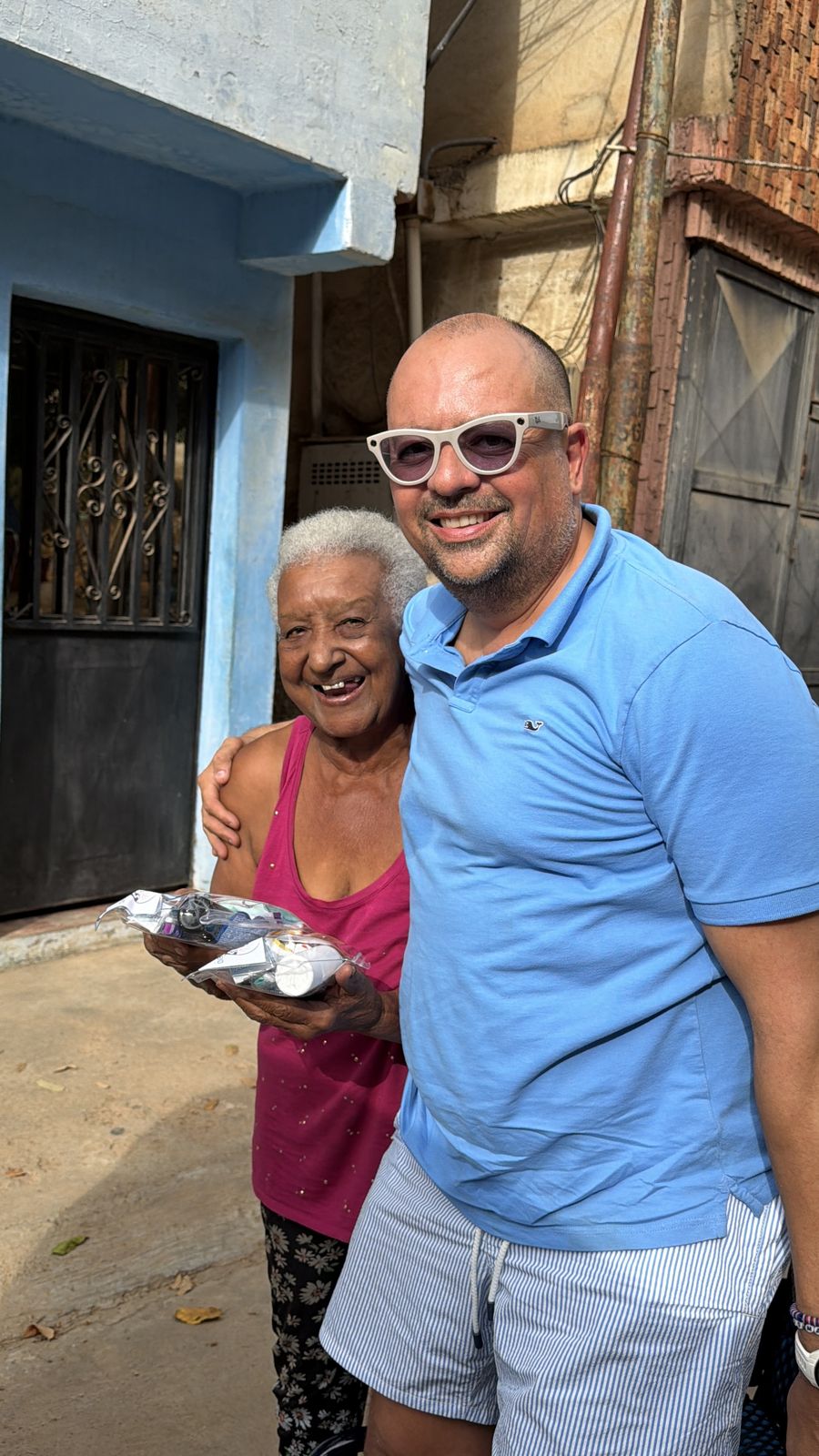 Un hombre con lentes de sol y camisa azul sonríe junto a una señora mayor que sostiene una bolsa de ayuda, frente a una casa.